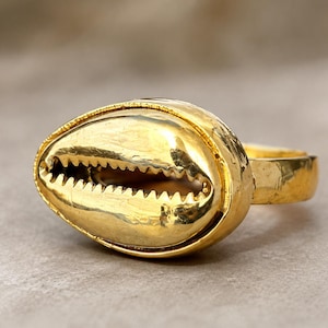 Pode incluir: Um anel de tom dourado com um design de concha de búzios como peça central. A concha tem um acabamento metálico liso com uma abertura horizontal central caracterizada por bordas serrilhadas e semelhantes a dentes ao longo da concha.