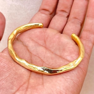 May include: A gold-toned cuff bracelet with an irregular, organic, and textured surface. The metal band features a continuous, undulating shape with a polished finish that reflects light across its uneven, bumpy contours and rounded edges.