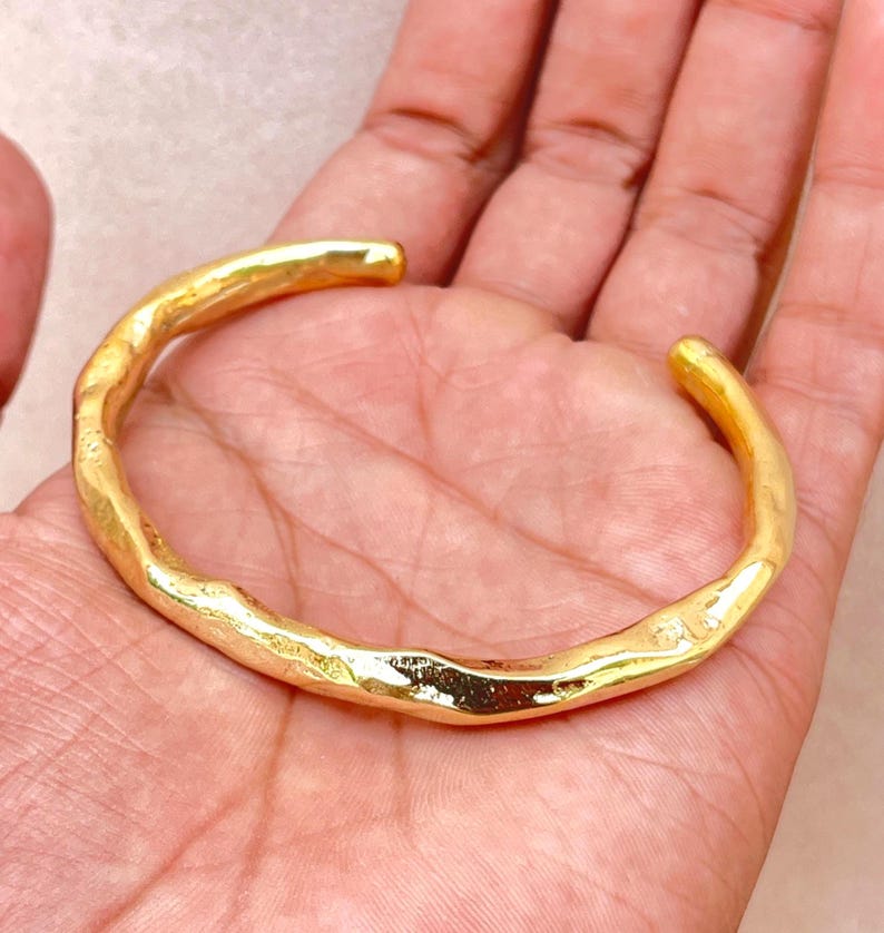 K&ouml;nnte beinhalten: Ein goldfarbenes Armband mit einer unregelm&auml;&szlig;igen, organischen und strukturierten Oberfl&auml;che. Das Metallband weist eine durchgehende, wellenf&ouml;rmige Form mit einer polierten Oberfl&auml;che auf, die das Licht auf den unebenen Konturen reflektiert.