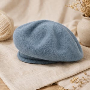 Op de afbeelding: Een zachte, stoffig blauwe gebreide baret met een afgeronde kroon en een geribbelde rand. De muts heeft een fijne, gelijkmatige textuur over het gehele oppervlak en toont een klassieke, ronde vorm die plat ligt. Het materiaal is pluizig gebreid.