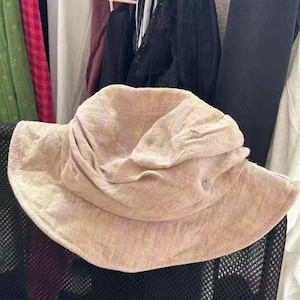 Puede incluir: Un sombrero de pescador de color rosa claro y amarillo con un acabado de tela texturizada. El sombrero presenta un ala ancha inclinada hacia abajo y una corona fruncida. Un pequeño botón metálico circular con el número 51 está en el lateral.