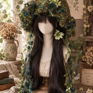 Skogsälva med mossblomma, handgjord fluffig stickad hinkhatt, älvhatt med skogsblommor och tofsar, huvudbonad i Goblincore Cottagecore