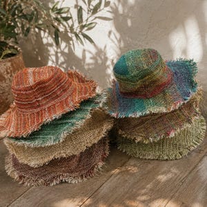 May include: A stack of bucket hats featuring frayed edges and textured woven fabric. The hats display a variety of colours including orange, green, brown, and a multi-coloured blend of blue, green, and purple, all with a visible coarse weave.