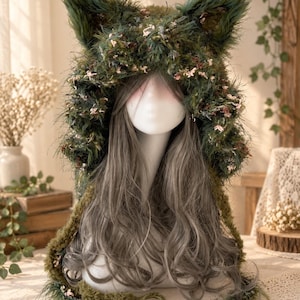 May include: A hooded accessory featuring pointed animal-like ears, crafted from a textured, shaggy dark green yarn interwoven with small pink and white floral accents. The hood drapes over a mannequin head with long, wavy, light grey hair.