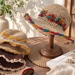 Könnte beinhalten: Ein beigefarbener Häkel-Sonnenhut mit einem regenbogenfarbenen Blumenband und Saum, neben einem kleineren beigen Hut mit gelbem Blumenband und einem dunkelbraunen Hut mit beigem Saum, alle mit aufwendigen, offenen Häkelmustern.