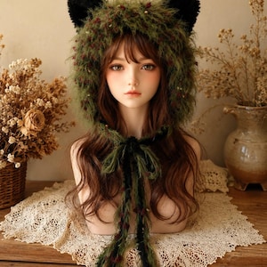 May include: A fuzzy olive green bonnet featuring two black cat ears on top and long, dangling ties. The bonnet is crafted from a textured, shaggy yarn with small dark red accents throughout, framing the face with a soft, layered appearance.