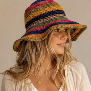Pode incluir: Um chapéu de crochê estilo bucket hat com listras horizontais em tons vibrantes de azul-marinho, verde-oliva, rosa brilhante e laranja. O chapéu possui uma copa arredondada e uma aba larga e flexível com acabamento em crochê verde-oliva.