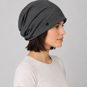 May include: A dark charcoal gray slouchy beanie hat made of a soft, textured knit fabric. The beanie features a relaxed, gathered fit that drapes toward the back of the head and includes a small, circular black button detail on the side.
