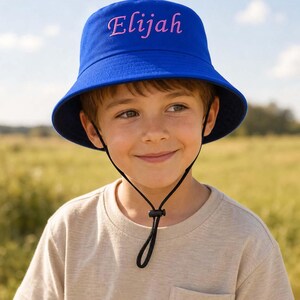 Peut inclure: Un chapeau bob bleu roi avec le prénom Elijah brodé en lettres cursives rose clair sur le devant. Le chapeau est doté d'une mentonnière noire réglable avec un cordon de serrage en plastique pour un ajustement sûr autour de la tête.