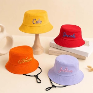 Peut inclure: Quatre chapeaux bob en jaune, rouge, orange et violet clair. Chaque chapeau présente des noms brodés : Cole en bleu, Jacob en bleu, Noah en jaune et Julian en rose. Deux chapeaux incluent des mentonnières noires avec des cordons réglables.