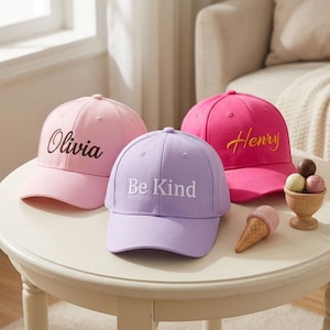 May include: Three baseball caps in light pink, lavender, and bright pink. The light pink cap features the name Olivia in dark brown script. The lavender cap has the text Be Kind in white. The bright pink cap features the name Henry in yellow.