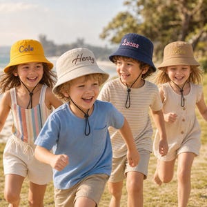 May include: Four bucket hats in yellow, beige, navy blue, and tan, each featuring an adjustable black chin strap and embroidered names: Cole, Henry, Lucas, and Finn. The hats are made of fabric and designed with a downward-sloping brim.