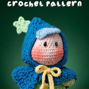 Op de afbeelding: Een gehaakte pop met een blauwe cape met capuchon en een lichtgroene ster aan de capuchon. De pop heeft een lichtroze gezicht met roze wangen, zwarte ogen en een geel lint met hartvormige uiteinden. De tekst luidt Twinkle Twinkle crochet pattern.