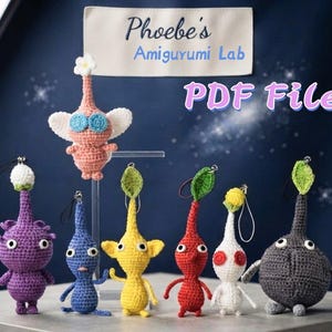 May include: A collection of seven crocheted amigurumi figures in various colors including purple, blue, yellow, red, white, and gray, each with a small loop for hanging. A sign above reads Phoebe's Amigurumi Lab and the text PDF File.