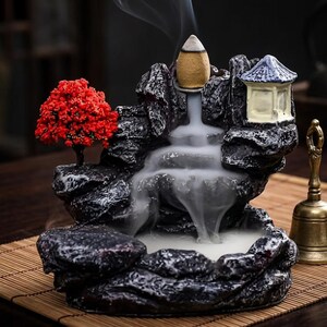 May include: A dark grey rock-shaped incense burner featuring a small red-leafed tree, a miniature pagoda structure, and a burning incense cone that releases white smoke flowing downwards like a waterfall into a basin, accompanied by a brass bell.