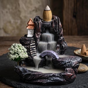 May include: A dark brown, rock-shaped incense burner featuring a miniature white pagoda, a small green tree, and a purple arched bridge. A lit incense cone sits at the top, releasing smoke that cascades down the sculpted tiers like a waterfall.