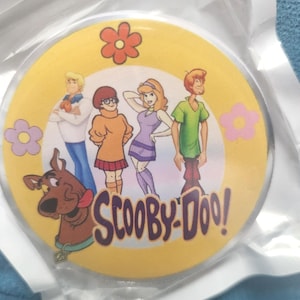 May include: A round pinback button featuring a yellow background with orange and pink flower graphics. The design displays the Scooby-Doo characters Fred, Velma, Daphne, Shaggy, and Scooby-Doo, with the purple text Scooby-Doo! printed at the bottom.