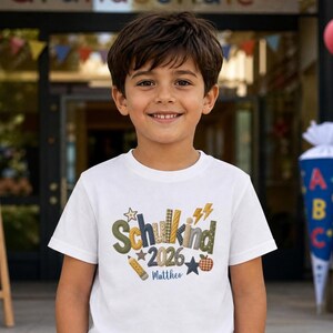 Könnte beinhalten: Ein weißes Kurzarm-T-Shirt mit dem Text Schulkind 2026 und dem Namen Mattheo. Die Grafik zeigt bunt gemusterte Buchstaben, einen Bleistift, einen Stern, einen Blitz und ein Apfelsymbol in den Farben Grün, Gelb und Braun.