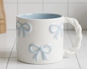 May include: A white ceramic mug featuring a repeating pattern of raised, light blue bows on the exterior. The interior of the mug is light blue, and it includes a white handle with a twisted, rope-like texture that extends from the side.