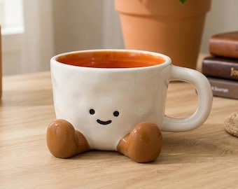 May include: A white ceramic mug with a textured, dimpled surface and a smooth handle. The mug features a simple black printed face with two dots for eyes and a curved line for a mouth. Two brown, rounded ceramic feet are attached to the base.
