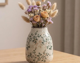 May include: A ceramic vase with a cream-colored base and a delicate green and pink floral vine pattern, filled with a dried flower arrangement featuring beige bunny tails, small purple statice, and soft orange rose buds in a rounded shape.