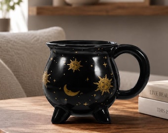 May include: A black ceramic mug shaped like a small cauldron with three short legs and a curved handle. The exterior features a gold-colored celestial pattern consisting of suns, crescent moons, and small stars scattered across the glossy surface.