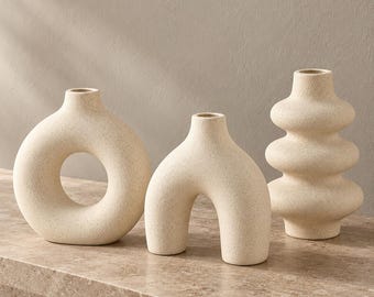 May include: A set of three off-white ceramic vases with a speckled, matte texture. The first vase is circular with a central opening, the second is arch-shaped with two legs, and the third is a stacked, multi-tiered vase with a narrow neck.