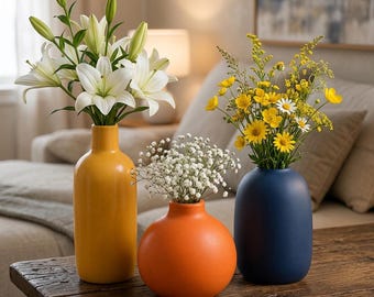 May include: Three ceramic vases in yellow, orange, and navy blue. The tall yellow vase holds white lilies, the round orange vase contains white baby's breath, and the cylindrical navy blue vase features yellow wildflowers and daisies.
