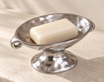 May include: A silver-toned metal soap dish with a hammered, textured finish and a pedestal base. The oval-shaped dish features small side handles and holds a single, rectangular, off-white bar of soap centered within the basin.