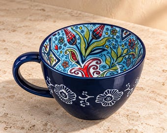 May include: A deep blue ceramic mug featuring a white floral pattern on the exterior and a vibrant interior design with red, green, and blue floral motifs. The mug has a curved handle and a wide, rounded bowl shape for holding beverages.