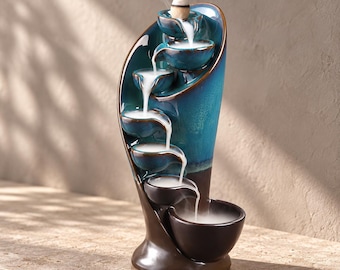 May include: A ceramic backflow incense burner featuring a vertical, curved design with a glossy teal upper section and a dark brown base. It has six stacked, bowl-shaped tiers that allow white incense smoke to cascade downward.