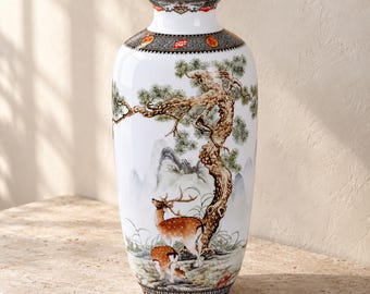 May include: A white ceramic vase featuring a detailed painted scene of two deer standing beneath a large pine tree with mountains in the background. The neck and base are decorated with intricate black, orange, and white geometric patterns.