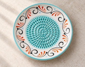 May include: A round ceramic grater plate featuring a central textured turquoise surface with raised spikes for grating. The rim is decorated with a repeating pattern of black swirling lines, orange leaf-like motifs, and small light blue dots.