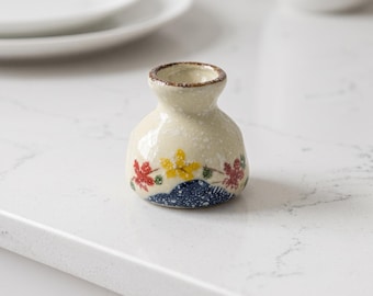 May include: A small, bulbous ceramic sake carafe with a cream-colored speckled glaze. The base features a dark blue mountain motif accented with painted red and yellow maple leaves. The rim is finished with a thin, dark brown painted edge.