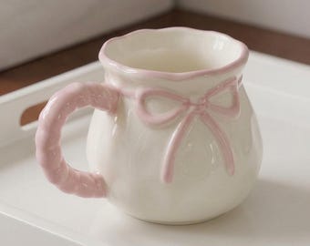 May include: A ceramic mug with a glossy cream-colored finish, featuring a scalloped rim and a raised pink bow design on the front. The mug has a textured, braided-style handle in pink, and the overall shape is rounded and bulbous.