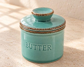 May include: A cylindrical ceramic butter crock in a light teal color with a matching lid. The word BUTTER is embossed on the front in capital letters. The rim of the lid and the base feature a dark brown speckled, rustic-style edge finish.