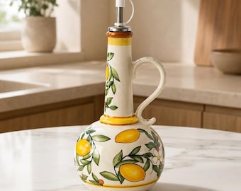 May include: A ceramic oil cruet with a round base and a tall, slender neck featuring a white, yellow, and green hand-painted lemon and leaf pattern. The vessel includes a curved handle and a silver-toned metal pour spout with a white plastic cap.