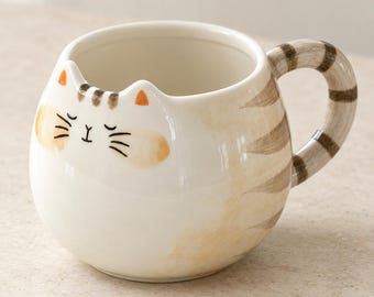 May include: A round ceramic mug shaped like a cat with a cream-colored body, light brown tabby stripes, and orange-tipped ears. The front features a painted face with closed eyes, a small nose, and whiskers. A striped handle extends from the side.