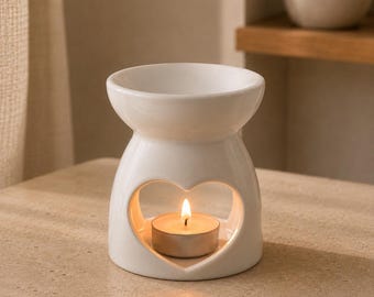 May include: A white ceramic oil burner featuring a heart-shaped cutout on the front. The burner has a wide bowl at the top for wax or oil and a hollow base that contains a small, lit tea light candle, casting a warm glow through the heart opening.