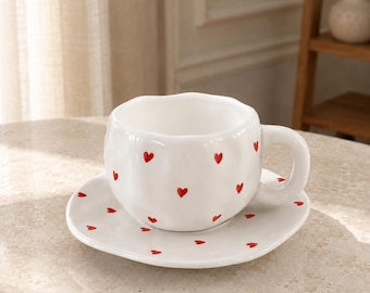 May include: A white ceramic teacup and matching saucer set featuring a hand-painted pattern of small red hearts. The cup has a rounded, slightly irregular shape with a thick handle, and the saucer has a matching wavy, organic edge design.