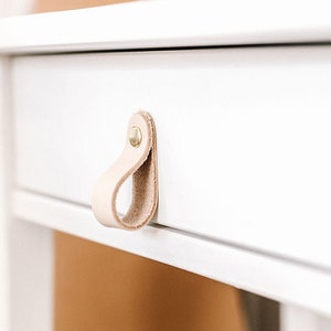 May include: Three white nightstands with drawers, each featuring a different leather pull. The pulls are in natural, dark blue, and light brown.