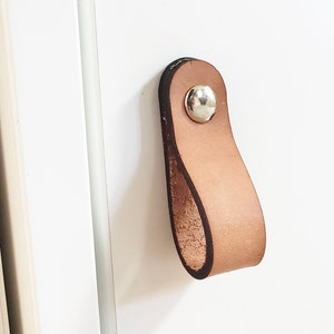 Könnte beinhalten: Ein hellbrauner Leder-Schrankgriff mit einer silbernen Niete, befestigt an einer weißen Schranktür. Der Griff hat eine Schlaufenform, und der Schrank ist aus Holz.