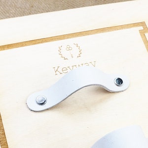 Könnte beinhalten: Zwei weiße Leder-Schubladengriffe mit silbernen Nieten, montiert auf einem Holzbrett mit dem Text "Keyway" und einem Logo eines Schlüssels mit einem Kranz darum.