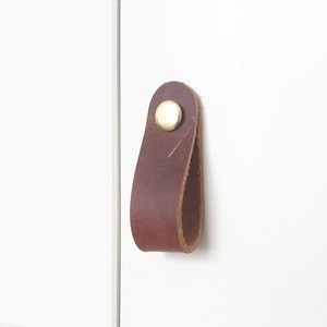 May include: A brown leather cabinet pull with a brass rivet. The pull is attached to a white door. This simple, modern hardware adds a touch of rustic charm to any cabinet or drawer.