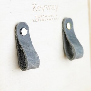 May include: Two gray leather drawer pulls with silver rivets. The pulls are mounted on a white wooden surface with the text "Keyway Hardware & Leatherworks" printed on it.