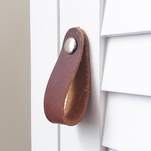 May include: A brown leather drawer pull with a silver rivet is attached to a white cabinet door. The pull is a loop shape, and the cabinet door has a shutter design. The background is a neutral color.