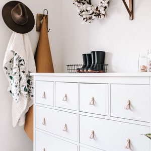 Puede incluir: Cómoda blanca con seis cajones y tiradores de cuero. La cómoda tiene una tapa blanca y está contra una pared blanca. Hay una corona de algodón en la pared sobre la cómoda.