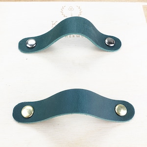 Puede incluir: Dos tiradores de armario de cuero verde oscuro con remaches plateados y dorados se muestran sobre una superficie de madera clara. Los tiradores tienen un diseño curvo. El fondo es una superficie de madera clara con un borde decorativo.