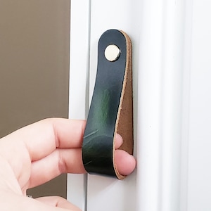 Può includere: Una maniglia per porta in pelle nera con una vite in ottone. La maniglia è fissata a una porta bianca.