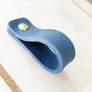 May include: A blue leather pull handle with a brass screw. The handle is attached to a light wood surface.
